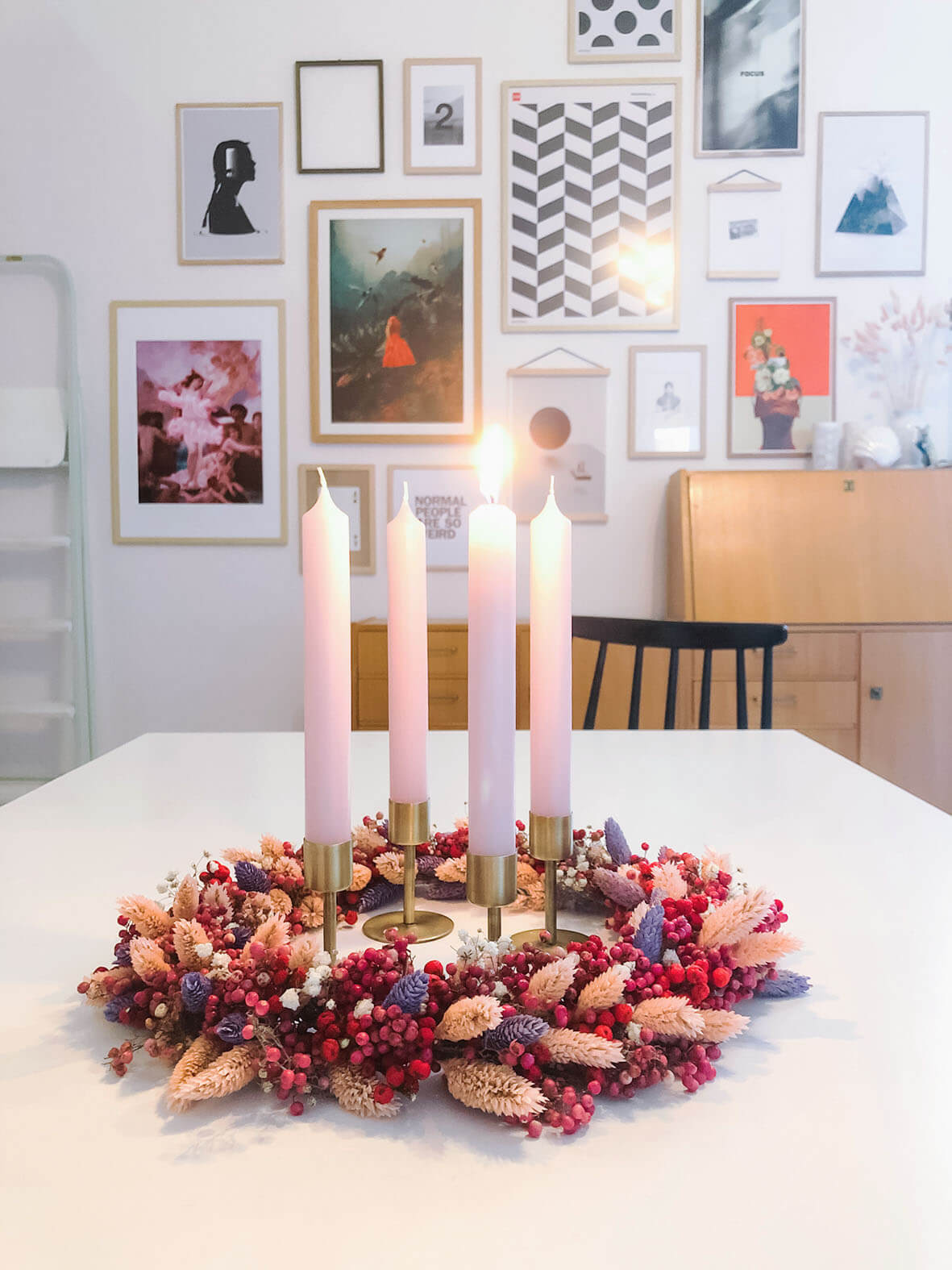 Der farbenfrohe Adventskranz liegt mittig auf dem weißen Esstisch. Die erste Kerze ist entzündet. Im Hintergrund ist eine Bilderwand zu sehen.