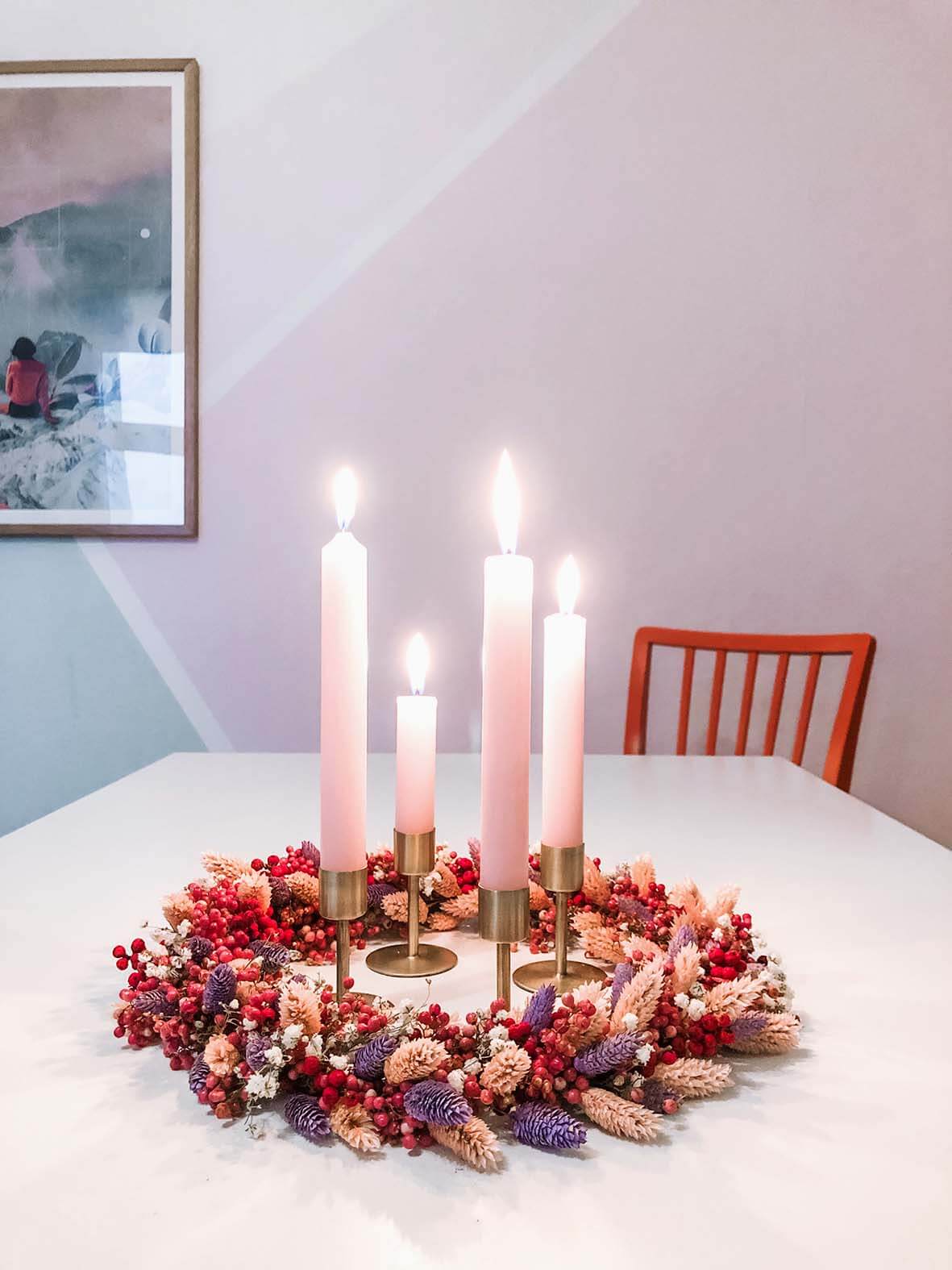 Der farbenfrohe Adventskranz liegt mittig auf dem weißen Esstisch. Alle vier Kerzen sind entzündet.
