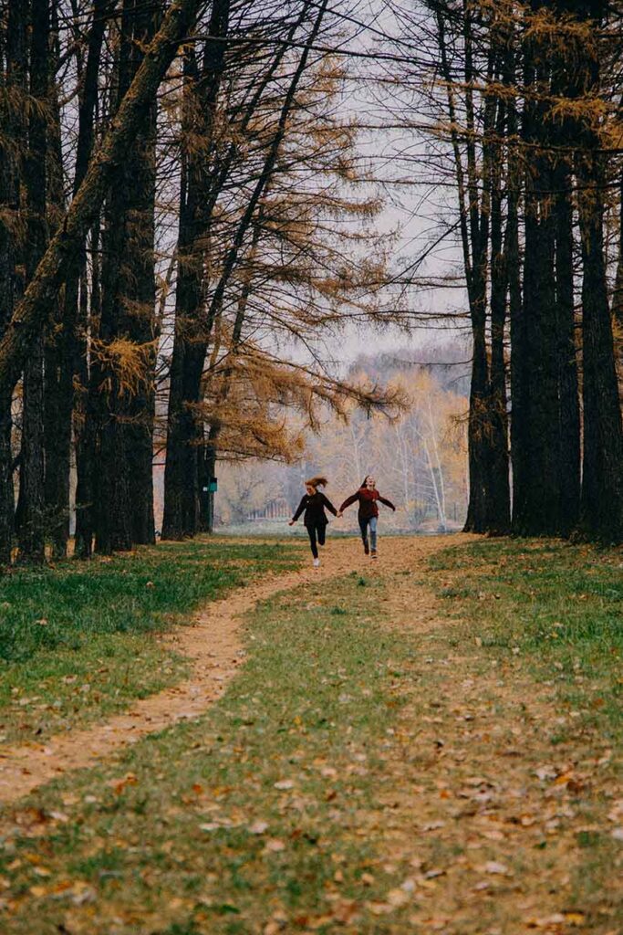 Zwei Freundinnen laufen durch einen Herbstwald zwischen hohen Bäumen