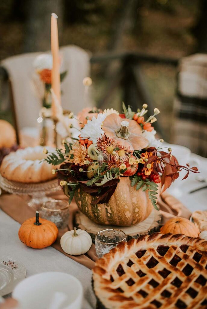 Kürbisvase mit Blumenarrangement als Tischdeko, umgeben von kleinen Kürbissen und Apfelkuchen