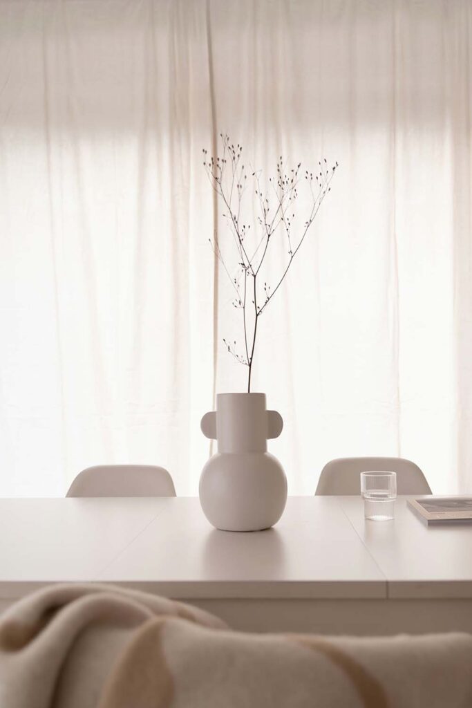 Schlichte Vase mit Zweigen auf weißem Tisch vor lichtdurchfluteten Vorhängen