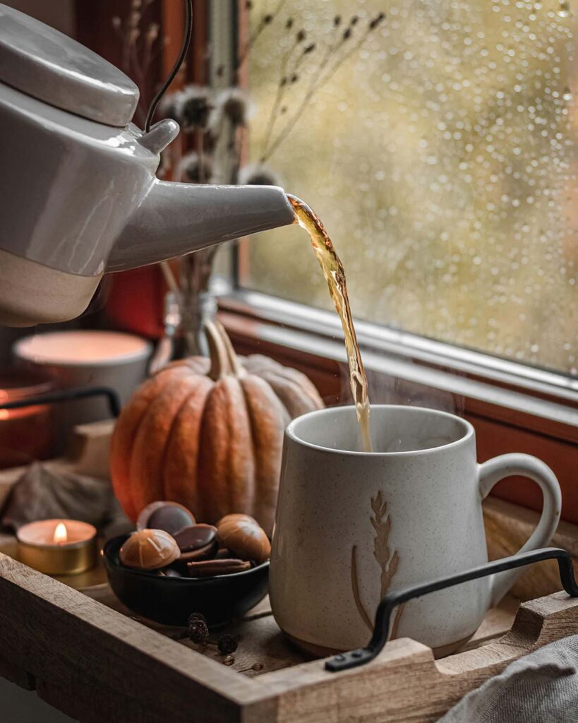 Teekanne gießt heißen Tee in Tasse, dekoriert mit Kürbis, Schokolade und Kerzen im Herbstambiente