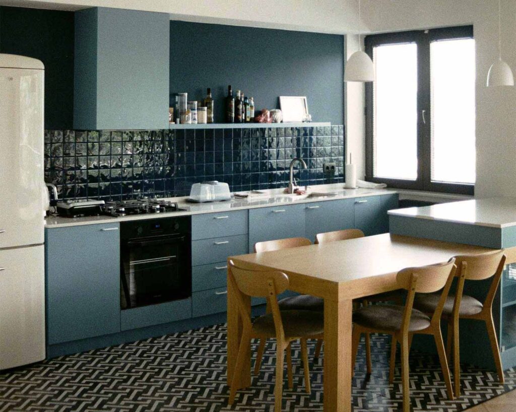 Kleine Küche in dunkelblau, mit dunkelblauem Fliesenspiegel. Davor ein großer grafisch gemusterter Teppich und ein Esstisch in hellem Holzton.