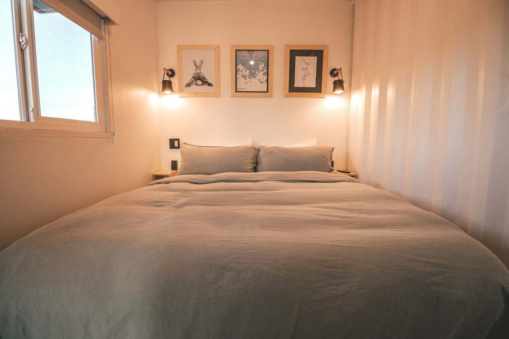 Kompaktes Schlafzimmer mit weicher Bettwäsche, warmem Licht und Bildern über dem Bett. Der Schlafbereich wird durch einen Vorhang vom Rest des Raums getrennt.