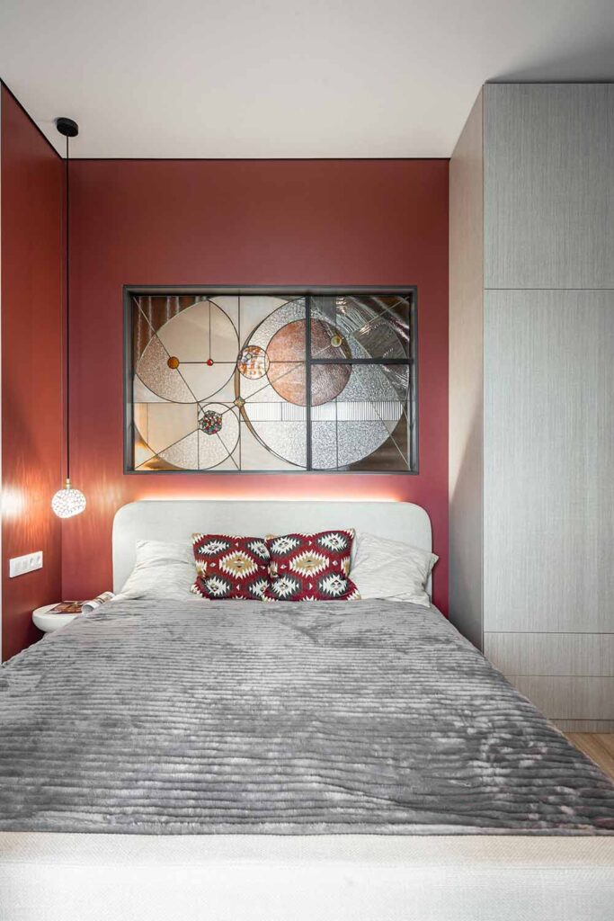 Kleines Schlafzimmer mit roter Wandfarbe und grafischem Kunstwerk aus Glas. Rotgemusterte Kissen auf dem Bett wiederholen die Wandfarbe