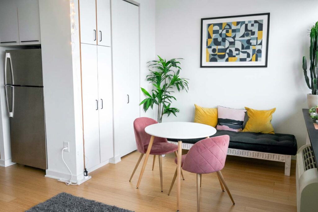 Kompakte Essecke in kleiner Wohnung mit rundem weißen Tisch, rosa Stühlen und kleiner Sitzbank mit gelben und rosa Kissen.
