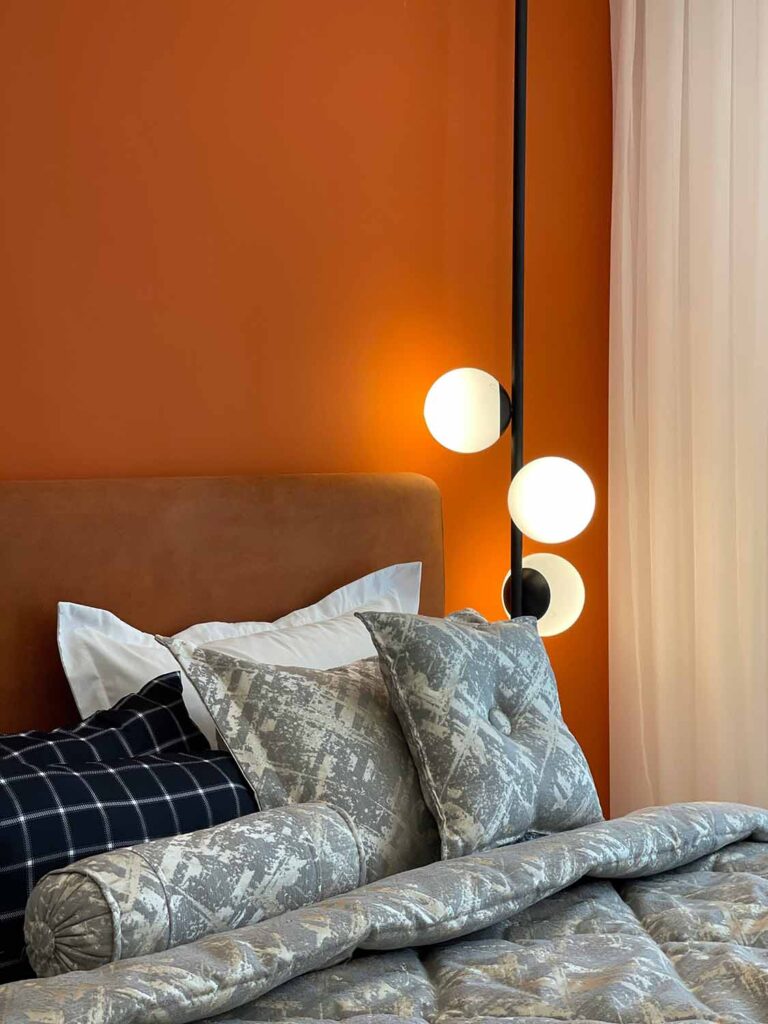 Ein Schlafzimmerdetail mit orangefarbener Wand, modernem Wandlicht und einem Bett mit gemusterten Kissen und gesteppter Tagesdecke. Helle Vorhänge rahmen das Bett seitlich ein.