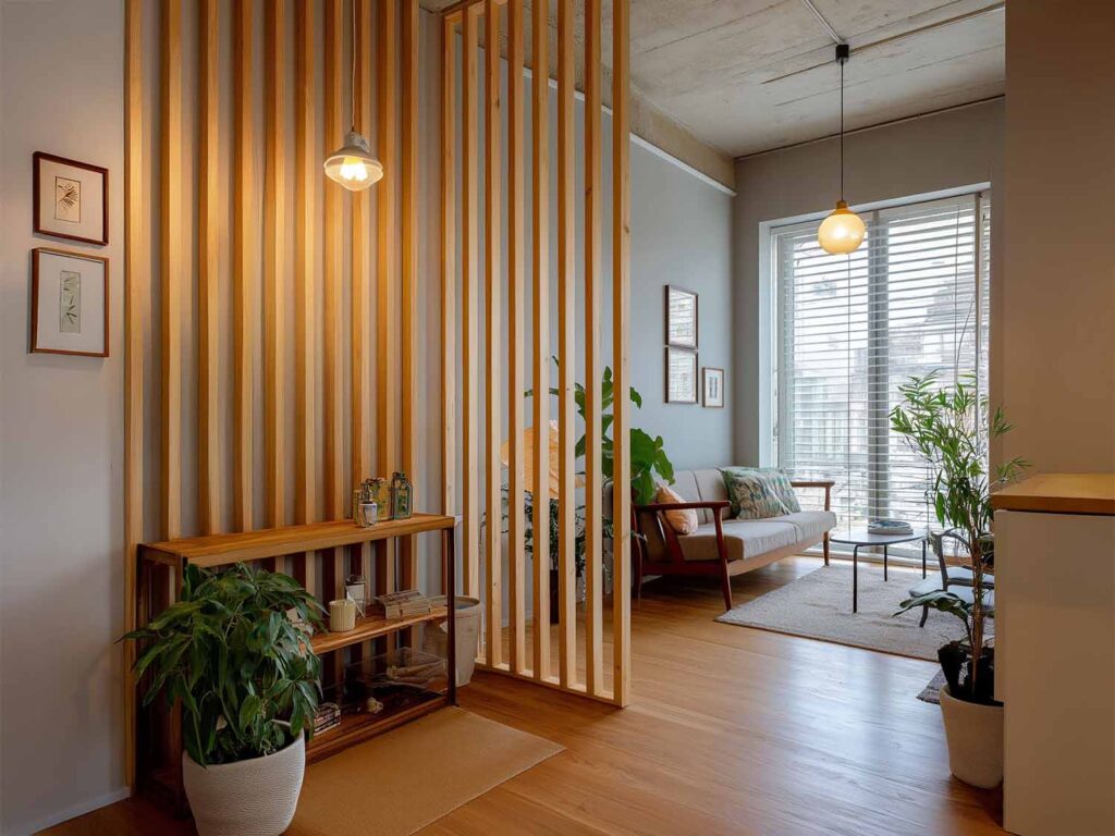 Basistipps für kleine Wohnungen: Ein luftiger Wohnbereich mit vertikalen Holzlamellen als Raumtrenner und viel Tageslicht. Pflanzen und Holzmöbel schaffen eine natürliche und warme Atmosphäre.