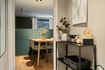 Ein Blick in einen kleinen Essbereich mit Holztisch und vier Stühlen vor einer grün gestrichenen Wand. Im Vordergrund steht ein schwarzes Regal mit Dekoobjekten, einer Vase und einer Lampe mit großer Deko-Birne.