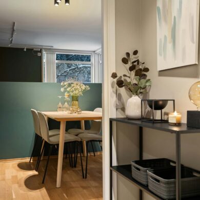 Ein Blick in einen kleinen Essbereich mit Holztisch und vier Stühlen vor einer grün gestrichenen Wand. Im Vordergrund steht ein schwarzes Regal mit Dekoobjekten, einer Vase und einer Lampe mit großer Deko-Birne.