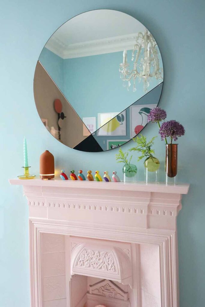 Ein runder Wandspiegel hängt über einem dekorativen Kaminsims vor einer hellblauen Wand. Auf dem Sims stehen Kerzen, kleine Figuren und Glasvasen mit Blumen.