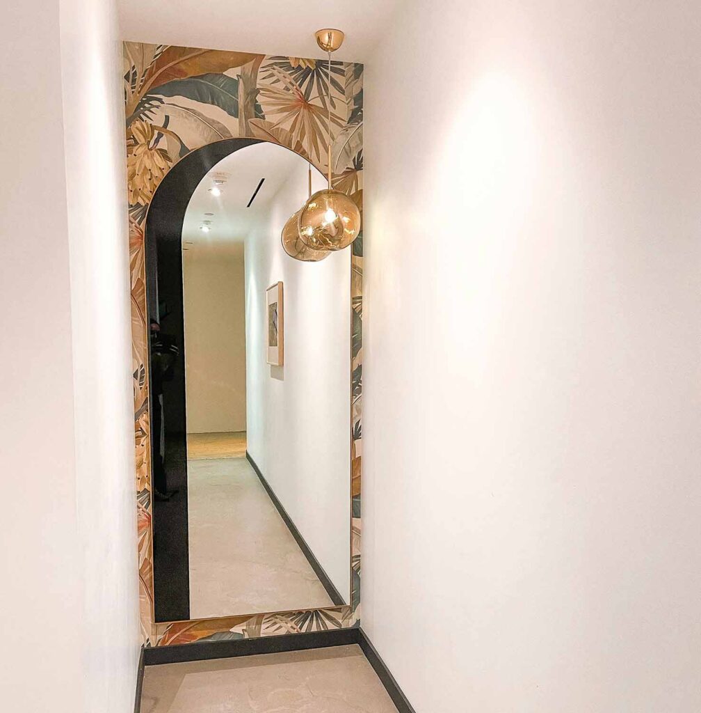 Ein schmaler Flur mit hohem Bogenspiegel, der den Flur reflektiert. Eine Pendelleuchte mit Glaskugel hängt vor dem Spiegel.
