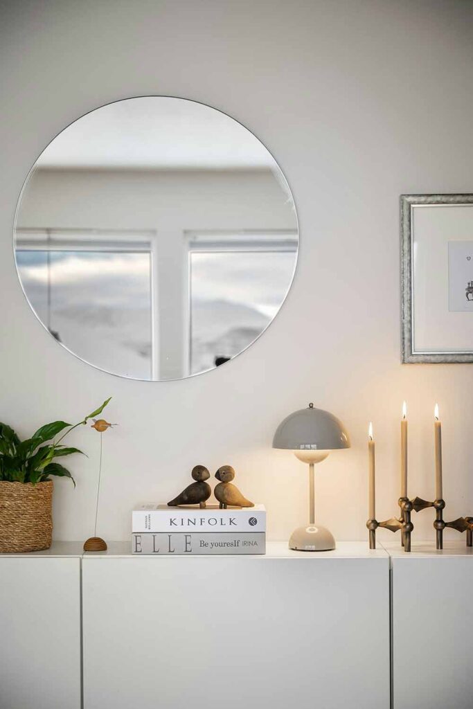 Ein runder Wandspiegel hängt über einem Sideboard mit Büchern, Kerzen und einer Tischlampe. Das Fenster gegenüber wird im Spiegel reflektiert.