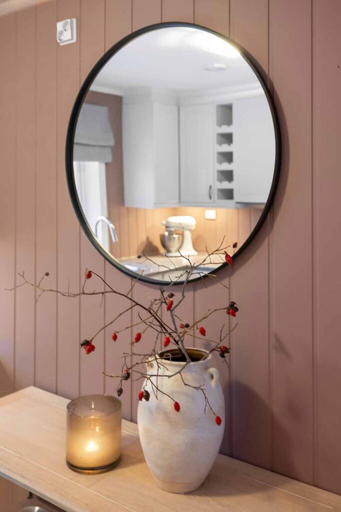 Ein runder Spiegel mit schwarzem Rahmen hängt über einer schmalen Konsole vor einer rosafarbenen Wand. Eine Kerze und eine Vase mit Zweigen stehen auf der Ablage.