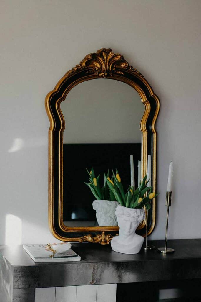 Ein Spiegel im Barockstil mit goldfarbenem Rahmen lehnt auf einem Sideboard und reflektiert einen Fernseher und Wanddeko. Auf dem Möbel stehen Kerzenhalter und eine Büste mit Tulpen.