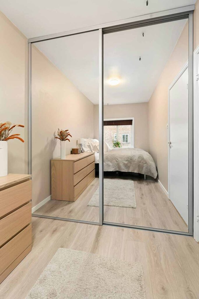 Ein Schlafzimmer mit Schiebetürenschrank, dessen Fronten als große Spiegel ausgeführt sind. Das Bett und eine Kommode spiegeln sich in den bodentiefen Spiegelflächen.