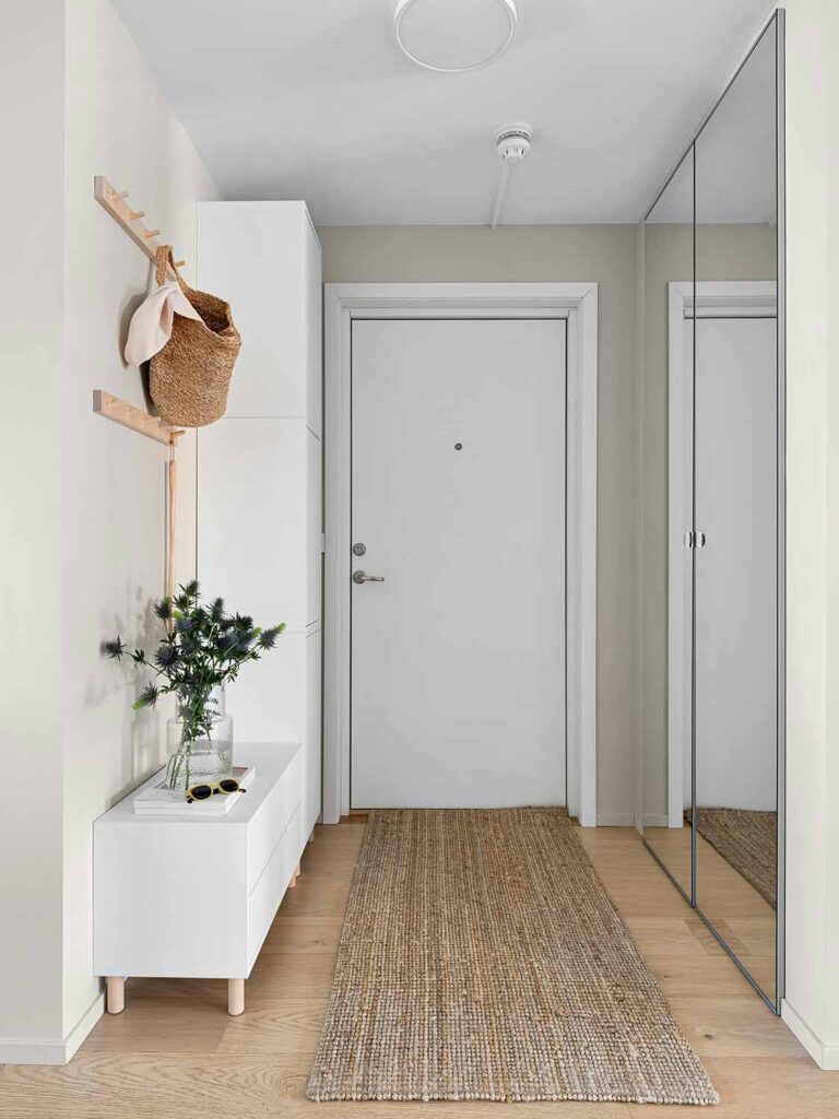 Ein heller Eingangsbereich mit weißer Tür, schmaler Sitzbank und großem Wandspiegel. Naturmaterialien und ein Läufer aus Jute ergänzen den ruhigen Look.