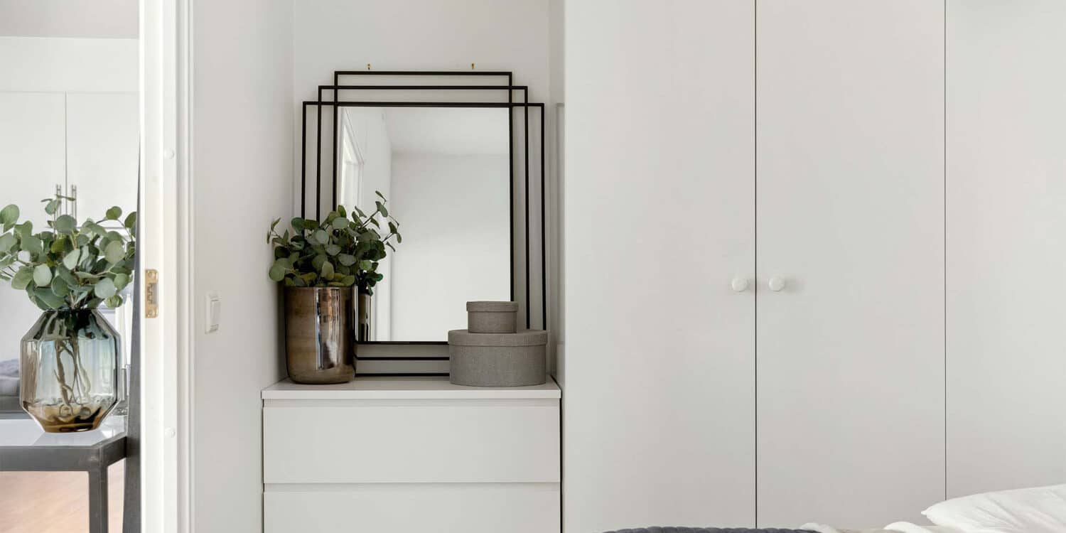 Ein Schlafzimmer mit weißer Kommode und einem rechteckigen Wandspiegel mit schwarzem Rahmen. Dekokörbe und eine Vase stehen auf der Kommode vor dem Spiegel.