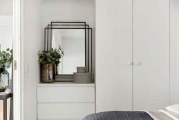 Ein Schlafzimmer mit weißer Kommode und einem rechteckigen Wandspiegel mit schwarzem Rahmen. Dekokörbe und eine Vase stehen auf der Kommode vor dem Spiegel.