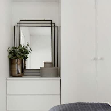 Ein Schlafzimmer mit weißer Kommode und einem rechteckigen Wandspiegel mit schwarzem Rahmen. Dekokörbe und eine Vase stehen auf der Kommode vor dem Spiegel.