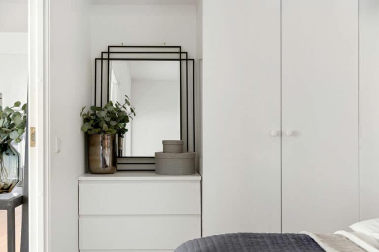 Ein Schlafzimmer mit weißer Kommode und einem rechteckigen Wandspiegel mit schwarzem Rahmen. Dekokörbe und eine Vase stehen auf der Kommode vor dem Spiegel.