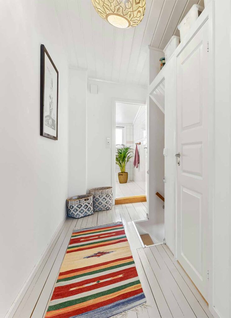 Schmaler weißer Flur mit Holzdecke, farbigem Läufer, Körben am Boden und offener Tür zum Bad