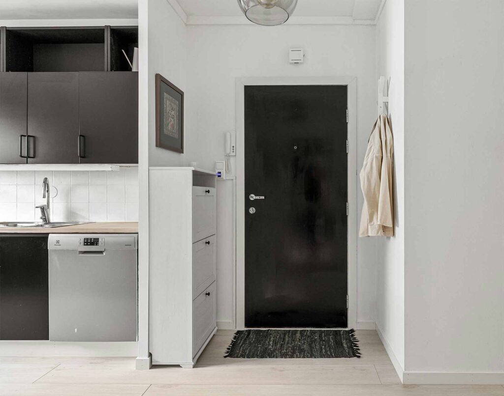 Schmaler Flur mit schwarzer Wohnungstür, kompaktem Schuhschrank und offener Küche im Hintergrund