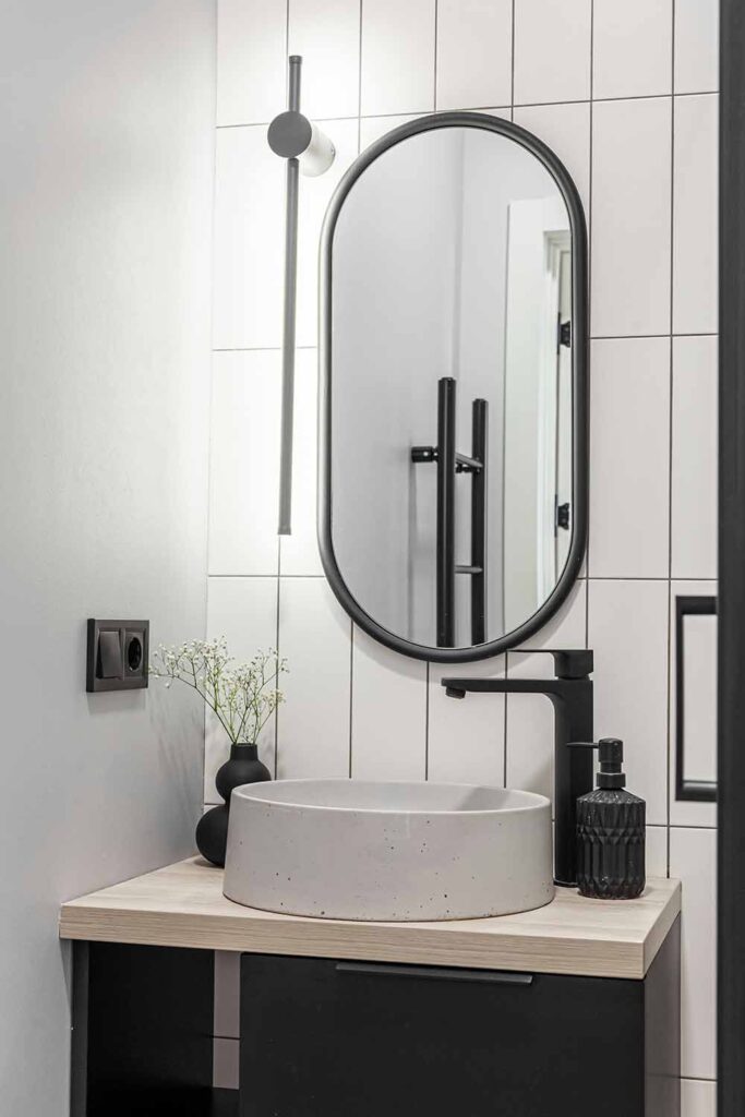 Moderner Waschplatz im kleinen Bad mit ovalem Spiegel, schwarzer Armatur und klaren Wandfliesen
