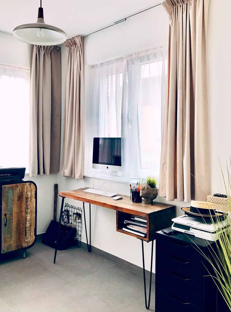 Schmaler Holzschreibtisch am Fenster mit Monitor, Vorhängen und Stauraum in kompakter Wohnraumsituation